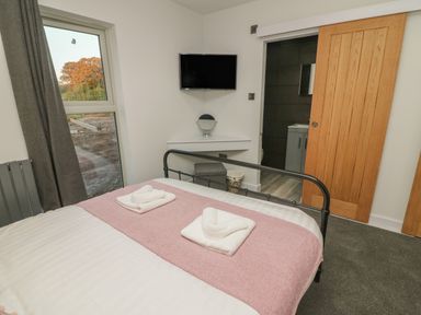 A bedroom with a bed with folded towels on a pink runner a wall-mounted tv a small vanity corner and a door leading to a bathroom at Box Tree Escapes in Wennington near Kirkby Lonsdale