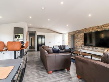 An open living room with brown leather sofas a wall-mounted television a bar counter with stools and a kitchen area at Box Tree Escapes Wennington near Kirkby Lonsdale