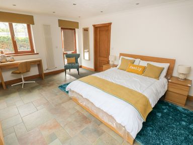 A bedroom with a bed with pillows and a throw a desk with a chair a blue armchair and two bedside tables with lamps at Oakland Cottage in Summerhill near Saundersfoot