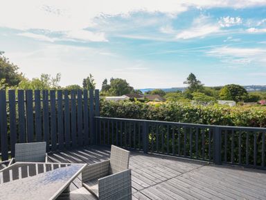 An outdoor deck with grey wicker chairs and a glass table overlooking greenery and distant houses at Oakland Cottage in Summerhill near Saundersfoot