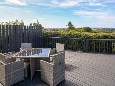 An outdoor deck with a glass table and wicker chairs overlooking trees and hills at Oakland Cottage Summerhill near Saundersfoot
