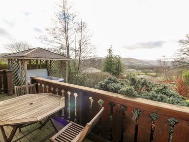 A wooden deck with table and chairs overlooking trees and hills with a gazebo covering a hot tub in the corner at Valley View in Rhayader