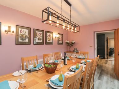A dining room with a wooden table set with plates napkins pears and wine a sideboard with flowers and framed pictures on the wall at Hazelnut Cottage in Windermere