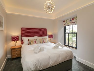 A bedroom with a double bed white bedding red headboard and lamps wooden bedside tables and a window with floral roman blind at Hazelnut Cottage in Windermere