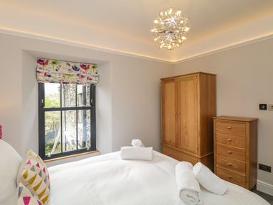 A bedroom with a bed with white linens and rolled towels near a window with floral blinds and wooden wardrobes at Hazelnut Cottage in Windermere