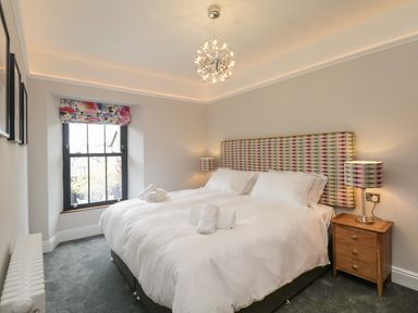 A bedroom with a large bed with white linens and patterned headboard two bedside tables with matching lamps and a window with floral blinds at Hazelnut Cottage in Windermere