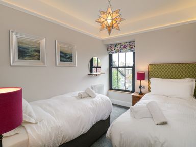 A bedroom with two single beds one with a green headboard and bedside tables with lamps and paintings on the wall at Hazelnut Cottage in Windermere