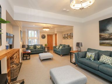 A living room with blue sofas, wooden side tables with lamps, two ottomans, a wall clock, and a TV at Hazelnut Cottage in Windermere