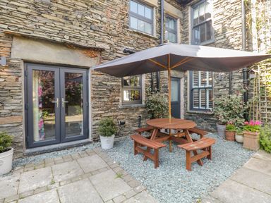 An outdoor patio area with a round wooden picnic table and umbrella surrounded by stone walls and potted plants at Hazelnut Cottage in Windermere
