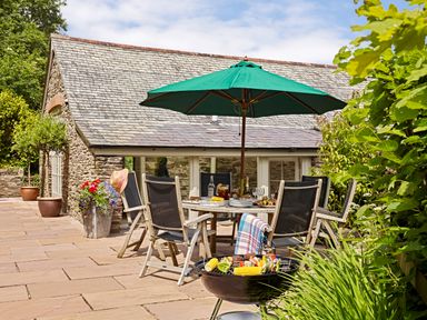 An outdoor patio area with a dining table and chairs under a green umbrella and a barbecue grill with food in front of a stone building at Garden House in Cornworthy