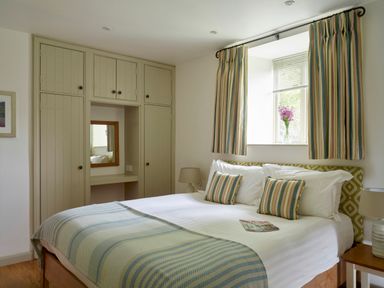 A bedroom with a bed with striped pillows and a blanket beside built in wardrobes and a window with striped curtains at Garden House in Cornworthy