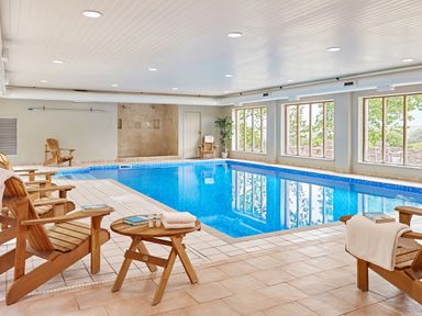 An indoor swimming pool with wooden chairs towels and books near windows at Garden House in Cornworthy
