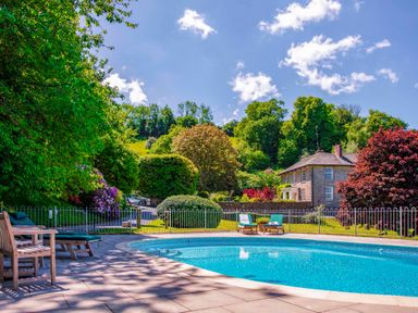 An outdoor swimming pool with lounge chairs and a stone house surrounded by trees at Garden House in Cornworthy