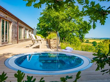 An outdoor patio with a round hot tub surrounded by wooden deck chairs and a wooden building with large windows at Garden House in Cornworthy