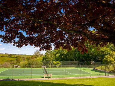 An outdoor tennis court with benches and a basketball hoop surrounded by trees and greenery at Garden House in Cornworthy