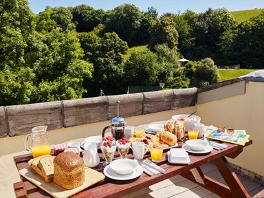 An outdoor wooden table set for breakfast with bread croissants orange juice and yogurt glasses at Haytor Cottage Cornworthy