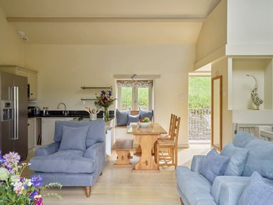 A living area with blue armchairs a wooden dining table with bench and chairs a kitchen with a refrigerator and flowers on the counter at Haytor Cottage in Cornworthy