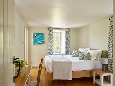 A bedroom with a wooden bed white bedding patterned pillows wooden side table with lamp and window with patterned curtains at Haytor Cottage in Cornworthy
