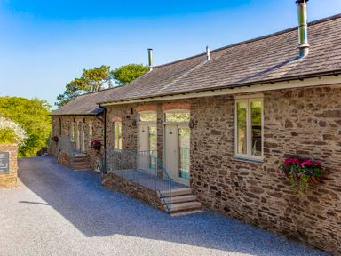 An exterior view of a stone building with multiple windows and doors along a gravel pathway with flower baskets attached to the wall at Haytor Cottage in Cornworthy