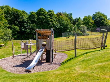 An outdoor playground with slides rope bridges and wood chips surrounded by grass and trees at Haytor Cottage in Cornworthy