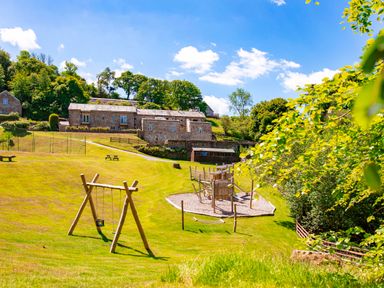A playground with swings a climbing structure and a hammock on a grassy hill with stone buildings and trees in the background at Haytor Cottage in Cornworthy