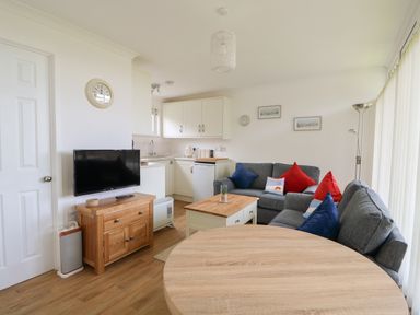 A living area with grey sofas with red and blue cushions a wooden coffee table and a round wooden dining table next to a kitchen area with white cabinets and a TV on a wooden stand at Pebbles in Cromer