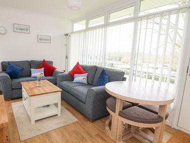 A living room with two grey sofas a wooden coffee table a round dining table with chairs and large windows with vertical blinds at Pebbles in Cromer