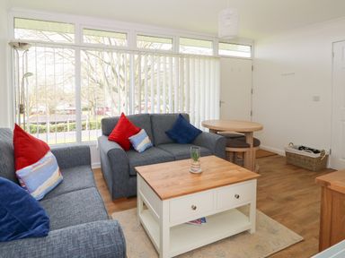 A living room with two grey sofas with red and blue cushions a wooden coffee table a round dining table with stools and large windows with vertical blinds at Pebbles in Cromer