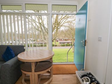 A living room area with a wooden table and cushioned stools next to a blue door with a view of trees and grass at Pebbles in Cromer