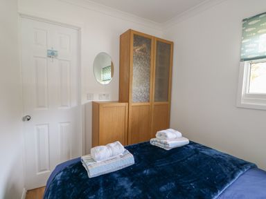 A bedroom with a bed covered in a dark blue blanket two sets of folded towels a wooden wardrobe with frosted glass doors a small cabinet a round wall mirror and a window with a fish pattern blind at Pebbles in Cromer