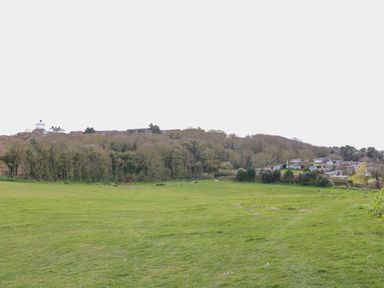 A large grassy field with a swing set near a wooded area and houses on a hill at Pebbles in Cromer