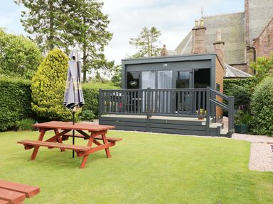 A garden with a wooden picnic table and umbrella in front of a small cabin with a deck at Cabin at the Tavern in Coupar Angus