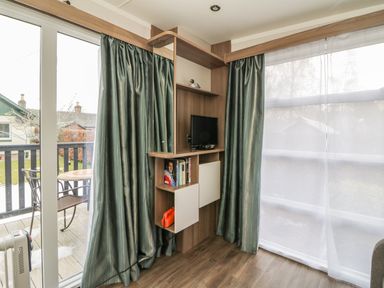 A corner in a room with wood floor shelves holding a small TV and books next to glass sliding doors and windows with curtains at Cabin at the Tavern in Coupar Angus