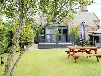 A garden with a picnic table and umbrella near a small decked cabin and a church building in the background at Cabin at the Tavern in Coupar Angus
