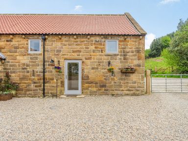 A stone building with two small windows a glass door hanging flower baskets gravel ground and a metal gate at Meadowsweet in Goathland