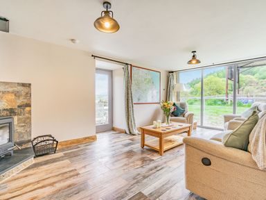 A living room with two armchairs a coffee table with flowers and candles a wood stove and large windows showing greenery outside at Meadowsweet in Goathland