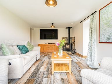 A living room with a beige sofa with cushions a wooden coffee table flowers a wall-mounted television a wooden cabinet a wood-burning stove and a curtain with a map at Meadowsweet Goathland
