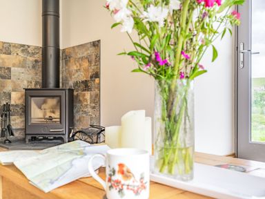 A wood burning stove with stone tiles behind it a vase with flowers candles a map and a mug on a wooden table at Meadowsweet in Goathland