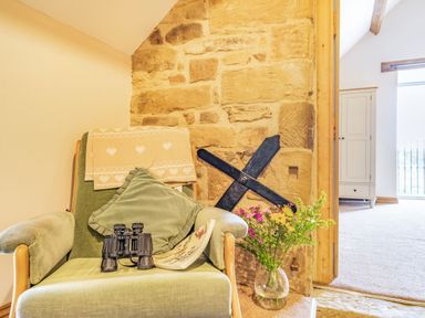 A green armchair with a blanket and cushion binoculars and a magazine on it beside a stone wall and a vase of flowers at Meadowsweet in Goathland