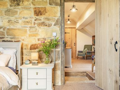 A bedroom with a white nightstand and flowers next to a stone wall and a wooden door leading to a hallway with an armchair at Meadowsweet in Goathland