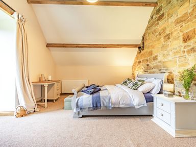 A bedroom with a bed, bedside table with a lamp and flowers, a desk, and exposed stone wall at Meadowsweet in Goathland