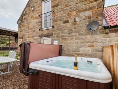 An outdoor hot tub with a bottle of prosecco and two glasses beside a stone wall and a table with chairs nearby at Meadowsweet in Goathland