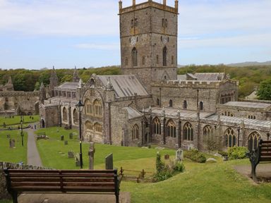 An old stone cathedral with a large tower surrounded by grass and gravestones with benches in the foreground at Pant y FFynnon in St Davids