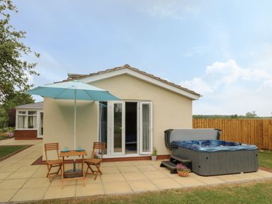 A patio with a wooden table and two chairs under a blue umbrella and a hot tub next to a house at Buttercups at Midway in Kington