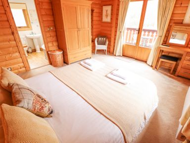 A bedroom with a bed covered in beige blankets two folded towels on the bed a wooden wardrobe a wicker basket a door leading to a bathroom a chair with a cushion a desk with a mirror and stool and glass doors with curtains and balcony outside at Great Leap in Louth