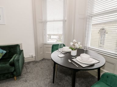 A dining table set for two with plates bowls glasses and cutlery near windows with blinds and green chairs and a green sofa with cushions at Emerald Suite in Barmouth