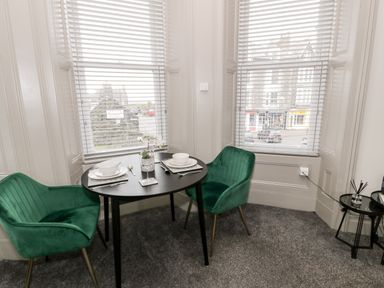 A dining area with a round table set for two and green chairs near two windows with blinds at Emerald Suite in Barmouth