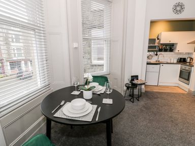 A dining area with a round table set for two next to windows and an open kitchen at Emerald Suite in Barmouth