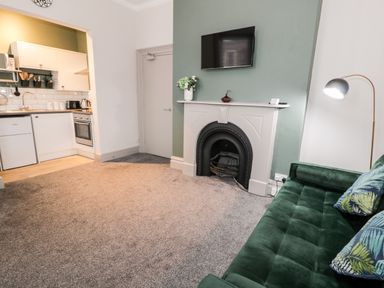 A living room with a green sofa two pillows a fireplace with a TV above and an open kitchen at Emerald Suite in Barmouth