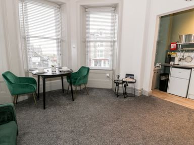 A dining area with two green chairs and a small round table near two windows with blinds and a partial view of a kitchen area at Emerald Suite in Barmouth